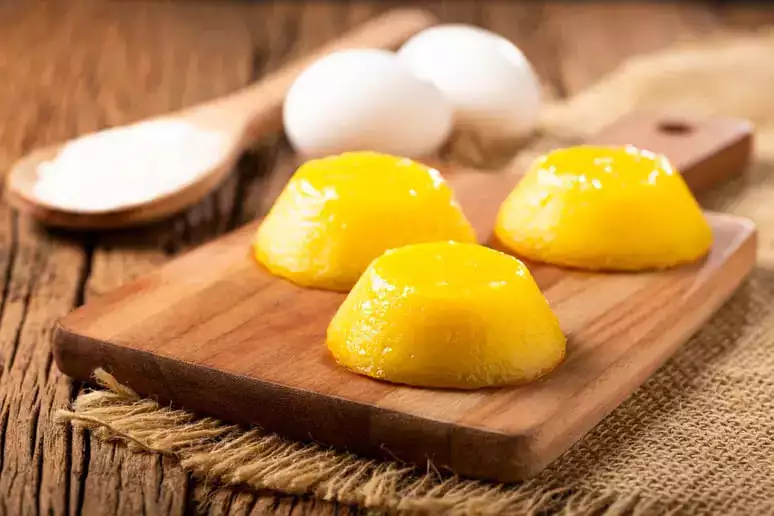 uma tábua com três quindins de goiaba com com queijo, ao lado dois ovos e uma colher de pau.