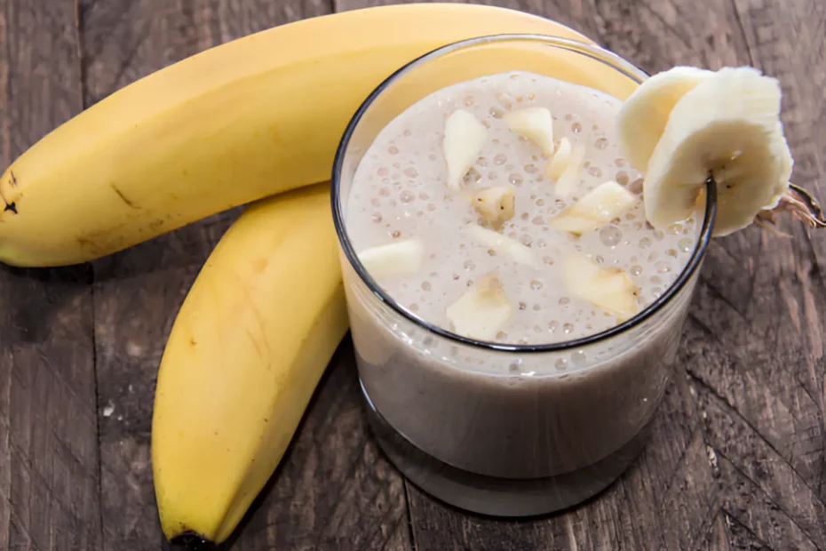 Um copo com uma vitamina de banana com creme de leite em cima de uma mesa com um cacho de banana ao lado para decorar.