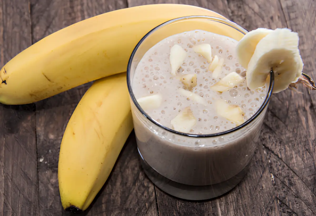 Um copo com uma vitamina de banana com creme de leite em cima de uma mesa com um cacho de banana ao lado para decorar.