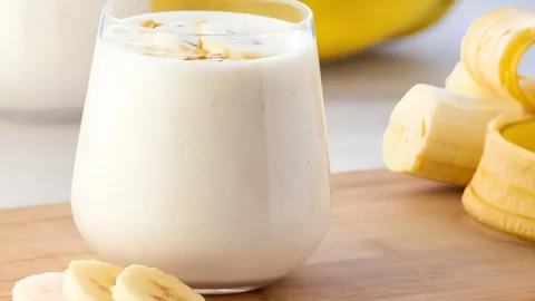 Um copo com uma vitamina de banana cremosa, ao lado uma banana picada para decorar a mesa.