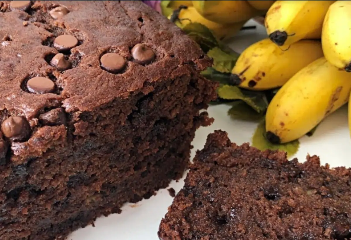 Um bolo de banana com gotas de chocolate com um cacho de bananas em cima da mesa para decorar.