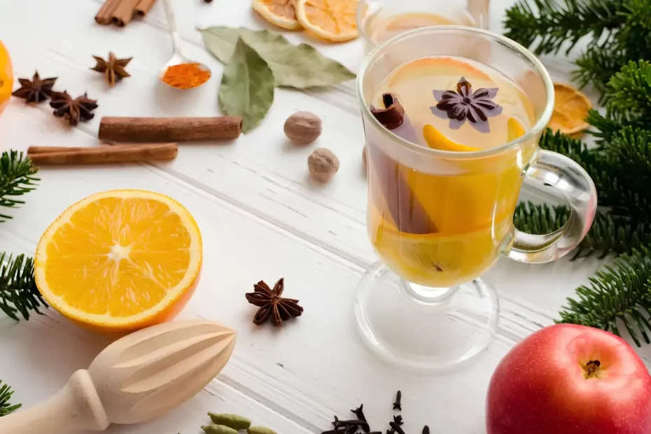 Uma taça com quentão de vinho branco e ao lado decorada com canela em pau, laranjas, folhas de loro.